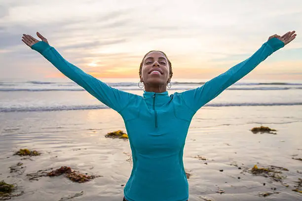 Celebration of Life. Beautiful woman if mixed race. Her arms raised up in the air. Her head is tilted back and eyes are close. She is in awe and smiling at the beauty of nature. Woman is expressing positivity, and feels grateful for life. She is very spiritual. Woman is at the beach with beautiful sunset in the background. 