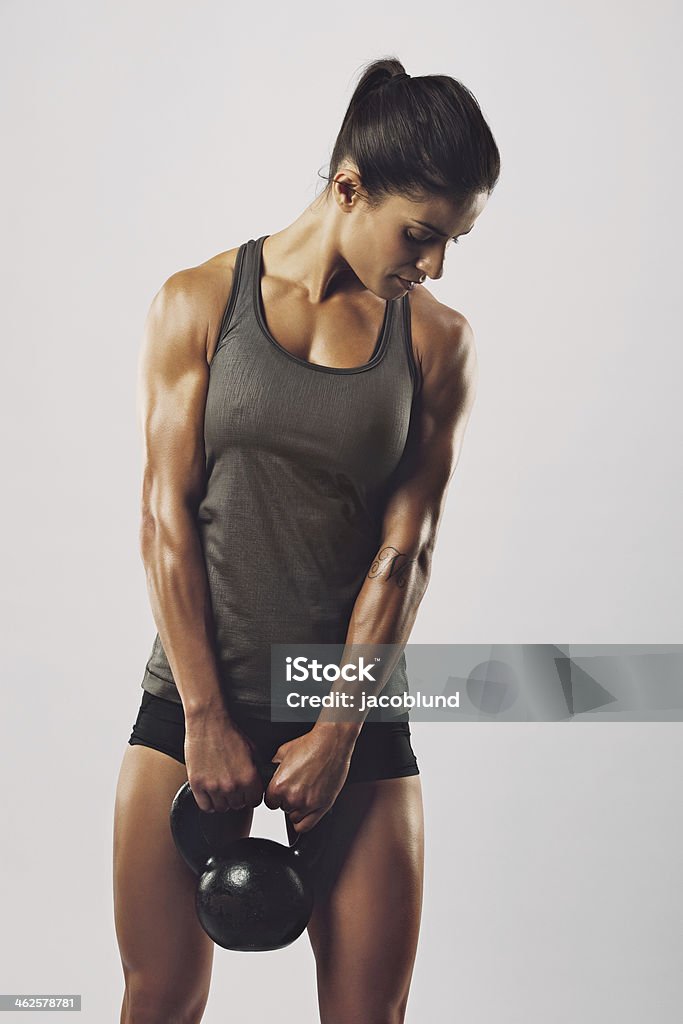 Fitness woman holding heavy kettle bell Fitness woman exercising gym holding kettle bell strength training biceps. Beautiful sweaty fitness instructor on grey background. Middle eastern female model with muscular fit and slim body. Cross Training Stock Photo