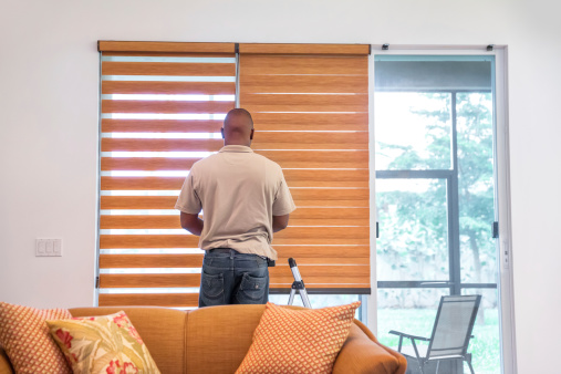A window treatment installer installs shades in a living room of a residential home.  RM