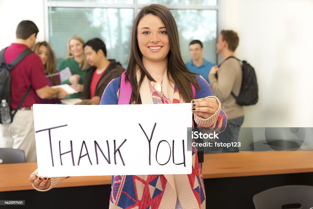 Education: Latin college student holds 'Thank you' sign. Female college student holds 'thank you' sign in appreciation. Classmates in background of classroom. Selective focus on sign. Talking Stock Photo