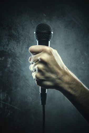 A mans hand grips a black mic, ready to emcee an event.  Would fit well as a representation of hip hop youth culture.  High contrast grunge styling to the image.  Vertical image with copy space.