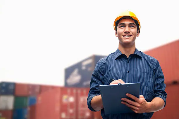 Photo of Smiling young engineer in a shipping yard examining cargo