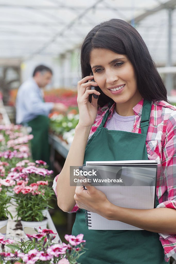 Garden center worker phoning in a greenhouse Brunette garden worker phoning in a greenhouse and holding a notepad 20-29 Years Stock Photo