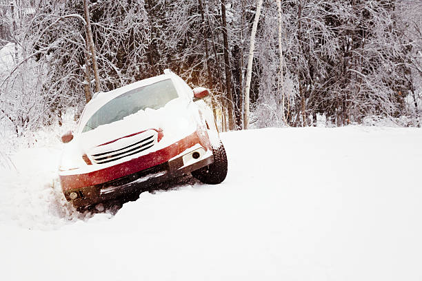 Winter snow accident featuring a car in a ditch on a snow-covered country road with the lights on. Falling snow visible, lots of snow on the car.