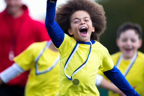 Young mixed race boy runs with his trophy held up looking at the camera. Team mates run behind celebrating.