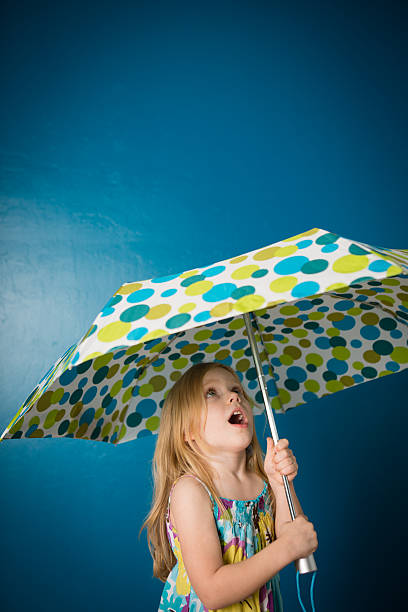 Color image of a surprised blond-haired little girl, standing under an umbrella she is holding. Includes blue background and copy space.