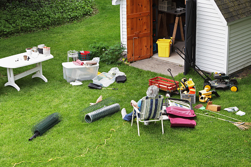 A mature, senior adult woman doing spring cleaning is resting in a folding chair after completely emptying the back yard shed of all sorts of outdoor household items, toys and gardening equipment.