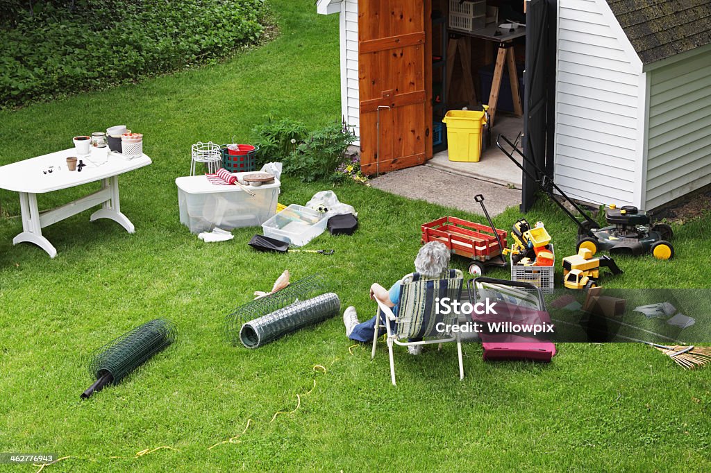 Overheard view of woman taking a break from spring cleaning A mature, senior adult woman doing spring cleaning is resting in a folding chair after completely emptying the back yard shed of all sorts of outdoor household items, toys and gardening equipment. Shed Stock Photo