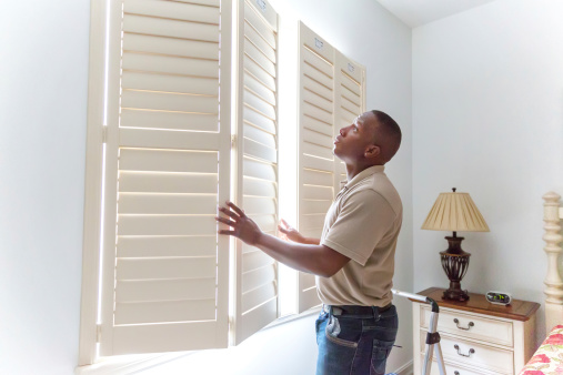 A window treatment installer installs wooden shutters in a residential home.  RM