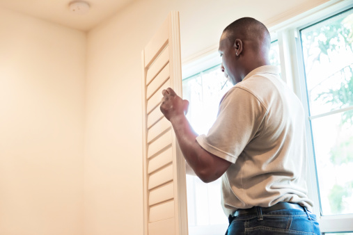 A window treatment installer carries a plantation shutter to the window of a residential home.  He is getting ready to install the shutter.  rr
