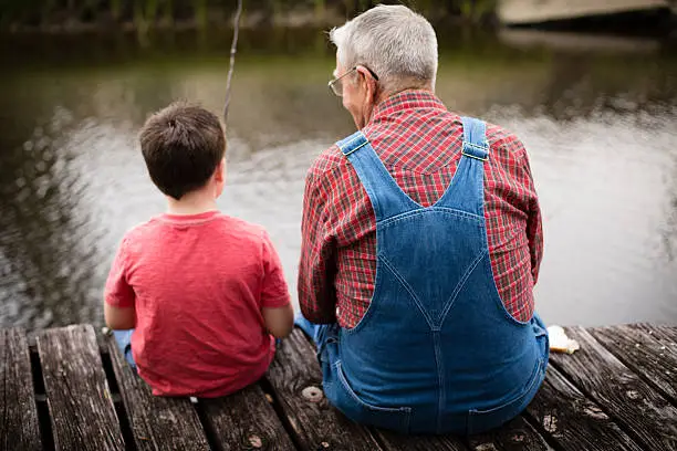 Color image of a young boy fishing with his grandfather while sitting on a dock.