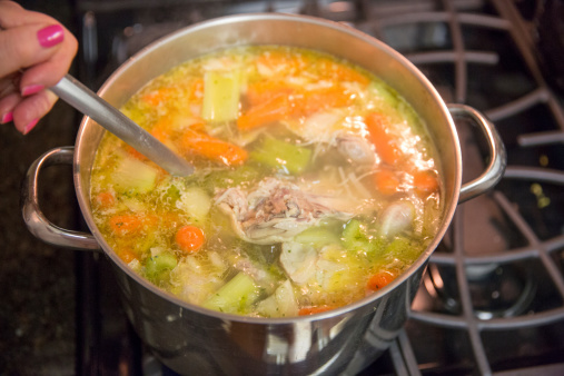 A pot of homemade chicken soup is cooking on the stove. Large pieces of chicken are in the soup.   RM