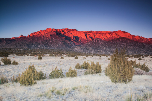 deep red alpenglow shines on the desert mountain ridges of peaks with a desert meadow of juniper stands far below.  such beautiful nature scenery can be found in the sandia mountains of albuquerque, new mexico.  horizontal wide angle composition.