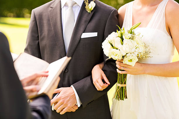 Midsection of bride and groom standing arm in arm with minister holding bible during wedding ceremony. Horizontal shot.