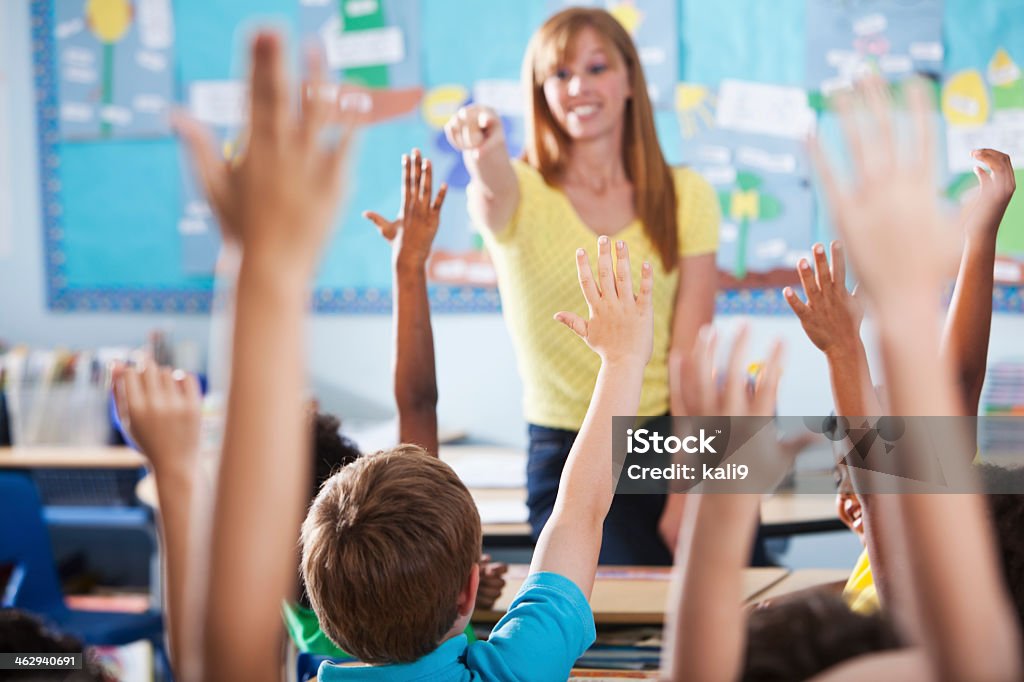 Elementary school class, raising hands Mature woman (40s) teaching class of multi-ethnic elementary school children. Focus on boy in blue shirt. Teacher Stock Photo