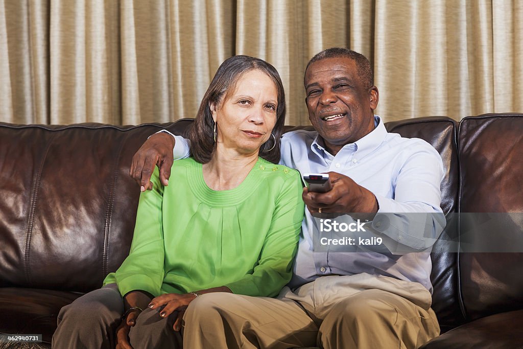 Senior African American couple watching TV on home on couch Senior African American couple (60s) sitting together side by side on couch with remote control, watching TV.  Man has his arm around woman resting his hand comfortably on her shoulder, holding remote with other hand, pointing it toward television, changing channels.  Husband is smiling, but wife has serious expression on her face. African Ethnicity Stock Photo