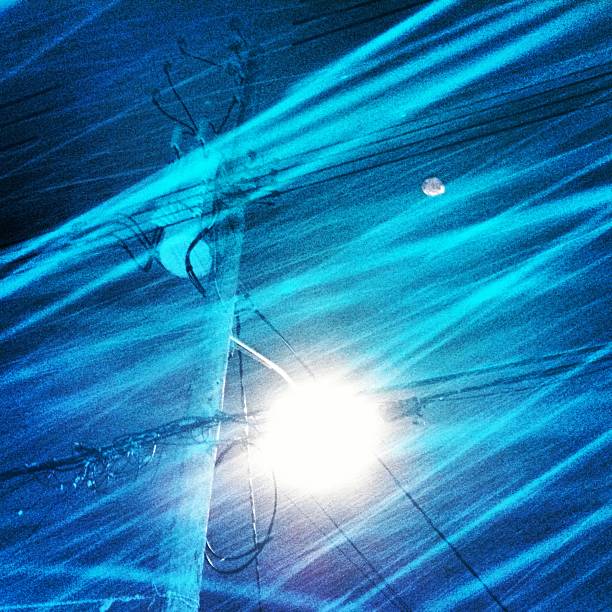 This is a square, mobilestock photograph of heavy snow falling during a blizzard at night. A flare is created by a street light on an electric pole.