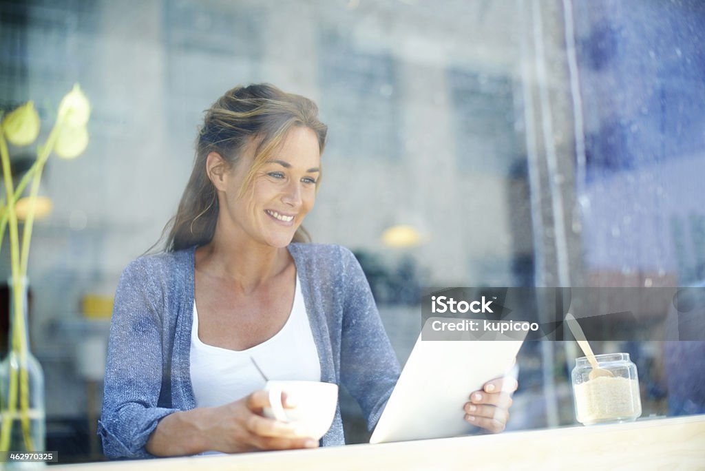 My favourite place to get some me time... A woman at a coffee shop sitting by the window smiling and looking at her touchscreen 30-34 Years Stock Photo