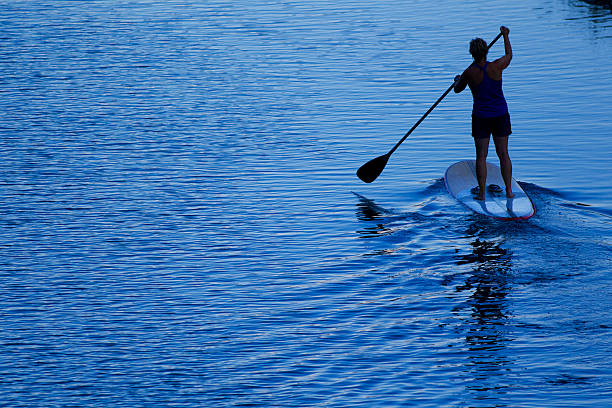 Young woman wearing a green life jacket and pink cap stands on a paddleboard on calm water under clear blue sky. The peaceful summer scene reflects outdoor recreation, balance, and healthy lifestyl