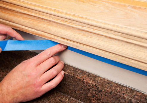 A painter is applying blue masking tape to protect an area of wall below oak window trim which is being refinished. The wall repainted after staining the trim, below the hand is a granite countertop.