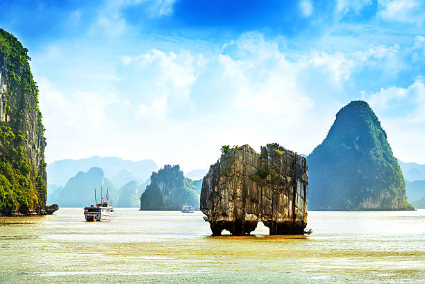 On a typical cloudy Halong Bay day, a tourist boat with sails aloft pauses at anchor amongst the spectacular limestone outcrops.  Only two unidentifiable people can be seen aboard.  Horizontal, all identification removed, copy space.