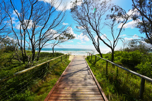 Wooden boardwalk beach access point at Broadbeach, Gold Coast, Queensland, Australia. Image shows wooden boardwalk and rails leading to the beach. Sky is blue and partially cloudy. Boardwalk has green vegetation and trees around it. Sandy beach with small surf.