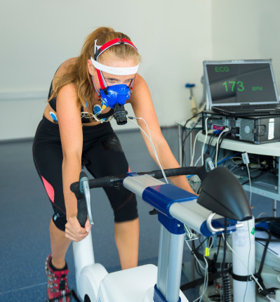 Female athlete having a VO2 test with a VO2 mask on her face, electrocardiogram pads attached, pulse rate 173 BPM, computer recording, indoor bicycle