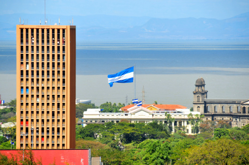 Managua, Nicaragua: Benjamin Zeledón tower, part of the parliament complex, former Banco de América tower (BAMER), lake Managua waterfront and Plaza de la Revolución / República with the Palace of Culture, flag, the Old Cathedral and the Presidential palace - seen from Loma de Tiscapa - Apoyeque volcanic complex mountains, Chiltepe Peninsula in the background - photo by M.Torres