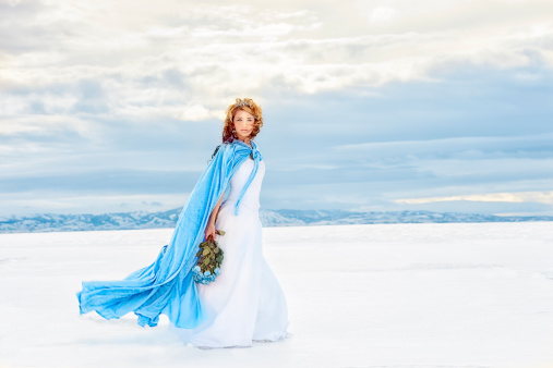 Beautiful Snow Queen Walking Across A Lake of Ice and Snow.  She wears her Tiara, her icy blue velvet cape, a snowy white dress and holds a frozen  bouquet of blue roses.  She is looking at the camera.  The wind is blowing her cape and hair as nature swirls all around her.  Cold Beauty.  Copy Space.