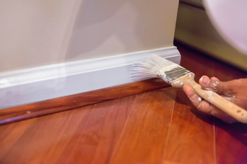 A house painter paints the baseboard of a residential home with white paint.  rr