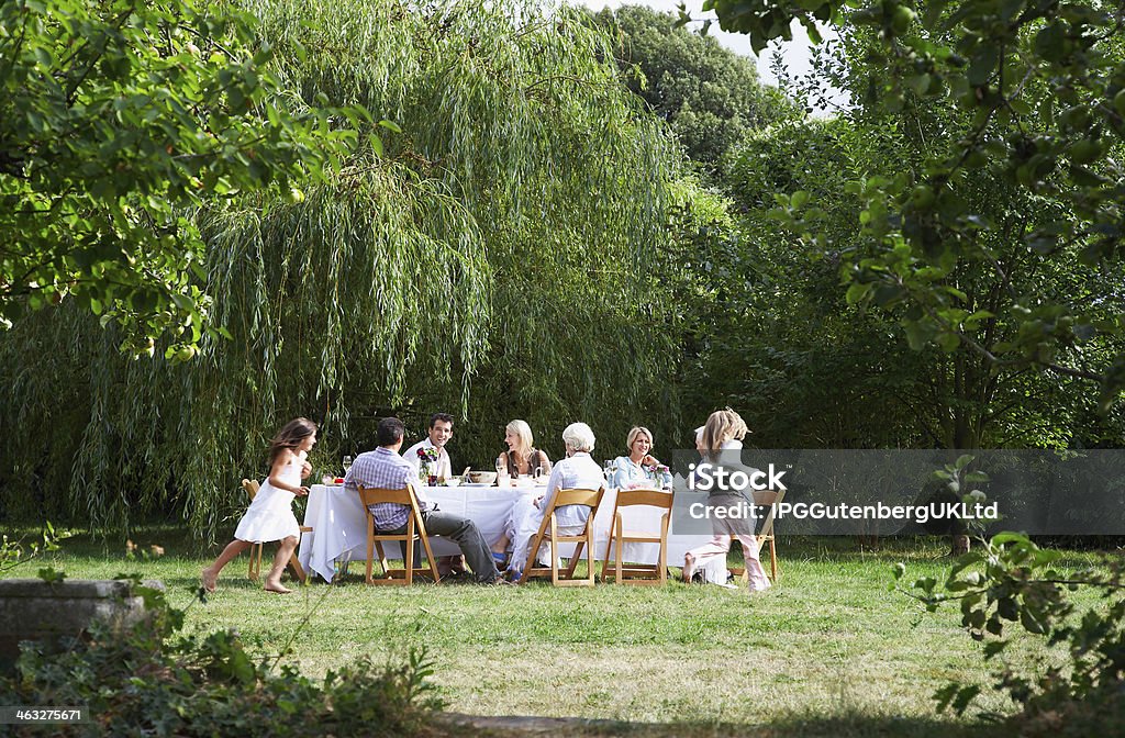 Happy Family Dining Together In Garden Happy family sitting at dining table while girls running around in garden Dining Stock Photo