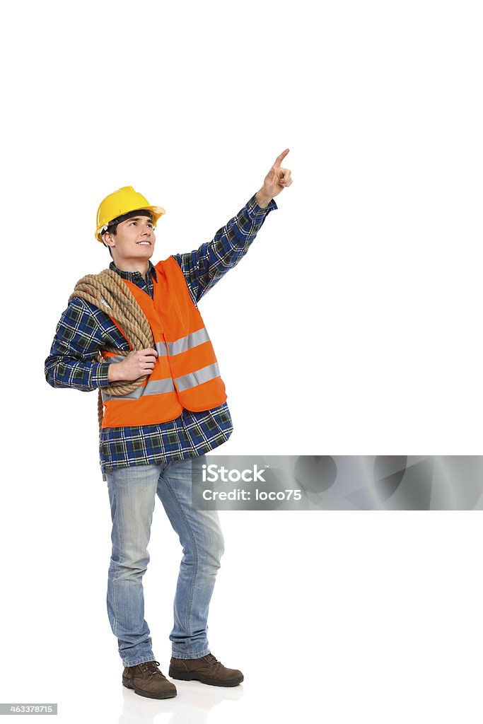 Construction worker holding rope and pointing up. Smiling construction worker in yellow helmet and reflected orange waistcoat pointing up. Full length studio shot isolated on white. Full Length Stock Photo