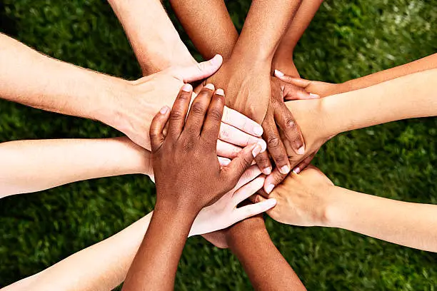 A pile of  multiracial hands are stacked in support or unity, against a background of grass. All for one and one for all! 