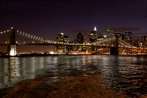 Brooklyn Bridge at Night, New York City. Brooklyn Bridge is the East River bridge connecting Manhattan and Brooklyn boroughs of New York City. East side of Downtown Manhattan is in background. The Statue of Liberty is visible at the left hand part of the image under the bridge.