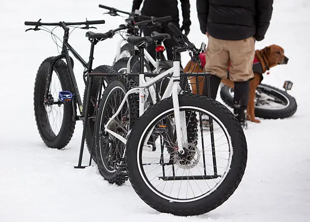 Fatbikes are sitting in a bike rack waiting to be ridden in a snow covered parking lot, while a couple of people and a dog hang out in the background.