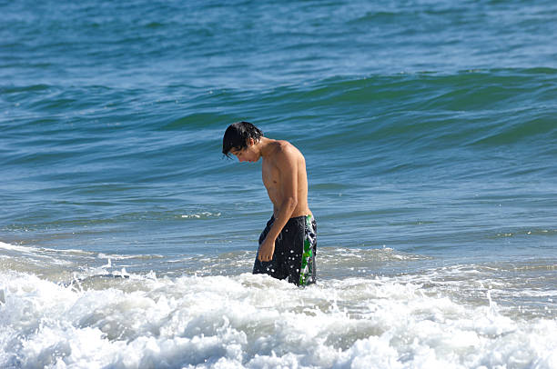 Teenage boy is enjoying a refreshing dip on beach in Normandy, France. He is lying on front, laughing as  he is being splashed by the breaking waves. Shot with Canon R5.