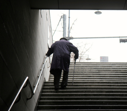 Back view of Elderly Man walking up a set of stairs, with the help of a walking stick, during winter.