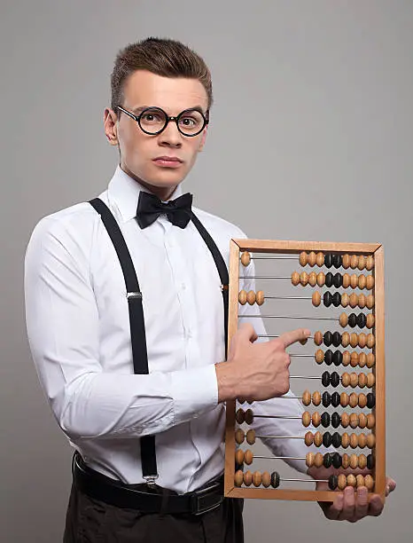 Serious young man in bow tie and suspenders holding abacus and pointing it while standing against grey background