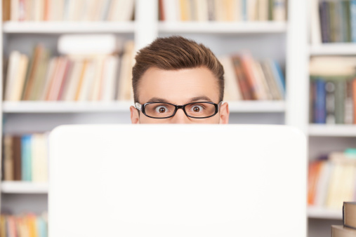 Shocked young nerd man in glasses looking out of computer while sitting at the library