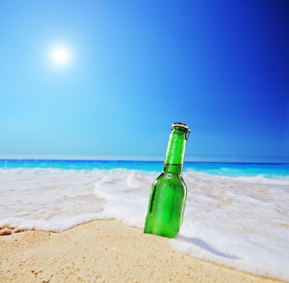 Beer bottle on a sandy beach with clear sky and wave, shot with a tilt and shift lens