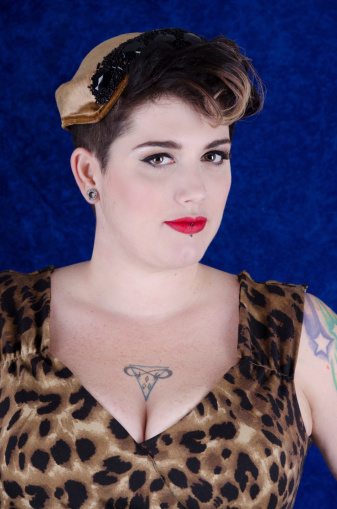 Vertical studio shot on blue of woman in tan and black vintage hat and leopard print dress with sweetheart neckline.