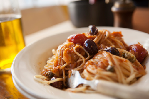 Puttanesca pasta, a traditional Italian spaghetti dish containing anchovy, garlic, capers, olives and tomatoes. Suggestion of beer and pepper grinder visible in the background.