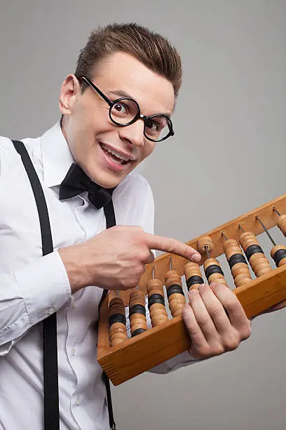 Low angle view of cheerful young man in bow tie and suspenders holding abacus and pointing it while standing against grey background