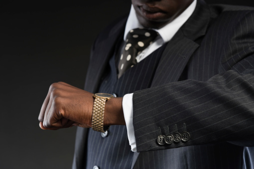 Retro african american gangster wearing striped suit and tie and black hat. Looking on his watch. Studio shot.