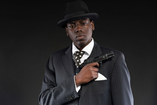 Retro african american mafia man wearing striped suit and tie and black hat. Holding a gun. Studio shot.