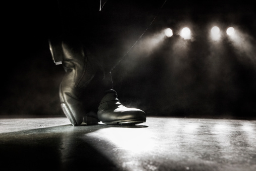 Tap dancer on an empty stage with lighting behind. Desaturated photo with visible noise and fog.