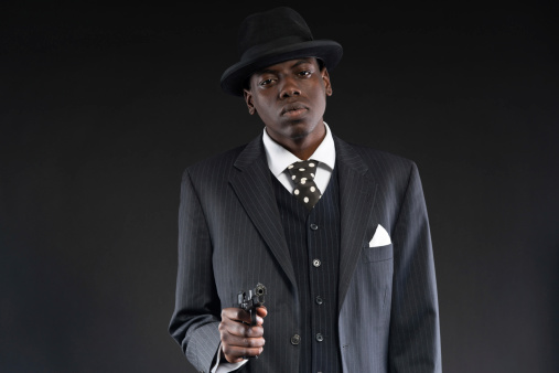 Retro african american mafia man wearing striped suit and tie and black hat. Shooting a with gun. Studio shot.