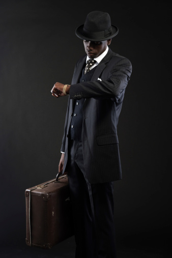 Retro african american traveller wearing striped suit and tie and black hat. Holding suitcase and looking on his watch. Studio shot.