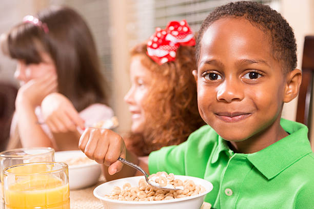 Food:  Three multi-ethnic children enjoying breakfast together. Three multi-ethnic friends have cereal and orange juice for breakfast.  African descent boy in foreground.  School or home setting.  boys bowl haircut stock pictures, royalty-free photos & images