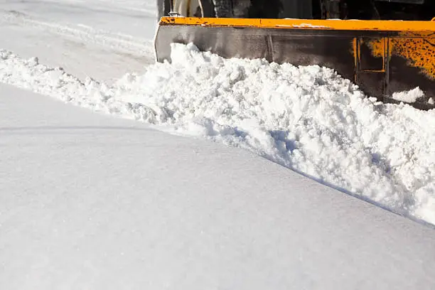 A large public works snowplow hydraulic wing section is plowing snow from a street. Focus is mid-frame, at 100% the plow blade is slightly soft.